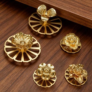 May include: A collection of five gold-toned metal cabinet knobs, each featuring a three-dimensional floral design centered on a flat, circular base with radiating cut-out spokes. The knobs vary in flower shape and size, displaying a polished finish.