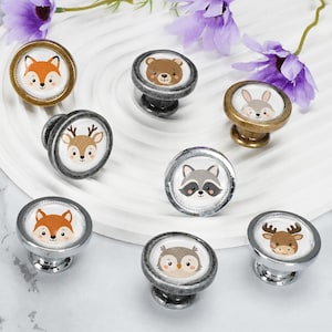 May include: A collection of eight round cabinet knobs featuring various animal illustrations including a fox, bear, rabbit, deer, raccoon, squirrel, owl, and moose. The knobs have metallic finishes in silver, bronze, and dark grey tones.