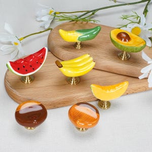 May include: A collection of six ceramic cabinet knobs shaped like various foods, including a watermelon slice, a banana bunch, an avocado half, a corn cob, a melon wedge, and two round nut-like shapes, all mounted on brass-colored metal bases.