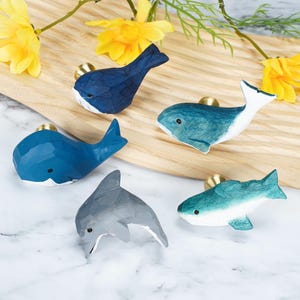 Op de afbeelding: Vijf gesneden houten ladeknoppen in de vorm van zeedieren, waaronder twee donkerblauwe walvissen, één blauwgroene walvis, één lichtblauwe walvis en één grijze dolfijn. Elke knop heeft een klein zwart oog en een goudkleurige metalen voet.