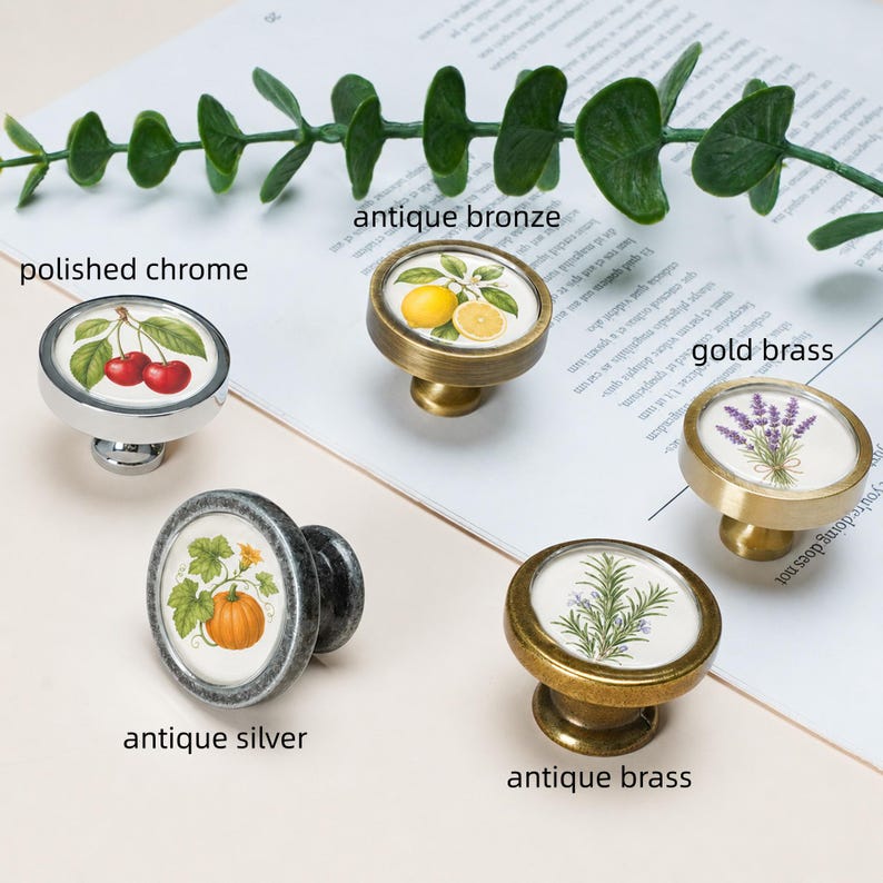 May include: Five round cabinet knobs with botanical illustrations. The knobs feature finishes labelled as polished chrome with cherries, antique bronze with lemons, gold brass with lavender, antique silver with a pumpkin, and antique brass with rosemary.
