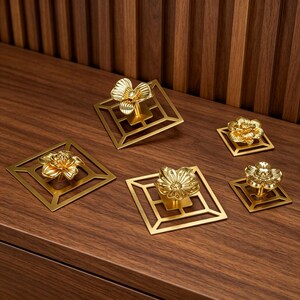 May include: A set of five gold-colored metal cabinet knobs, each featuring a three-dimensional flower shape mounted on a flat, square, open-frame base. The knobs vary in size, with some larger and some smaller, all displaying a polished finish.