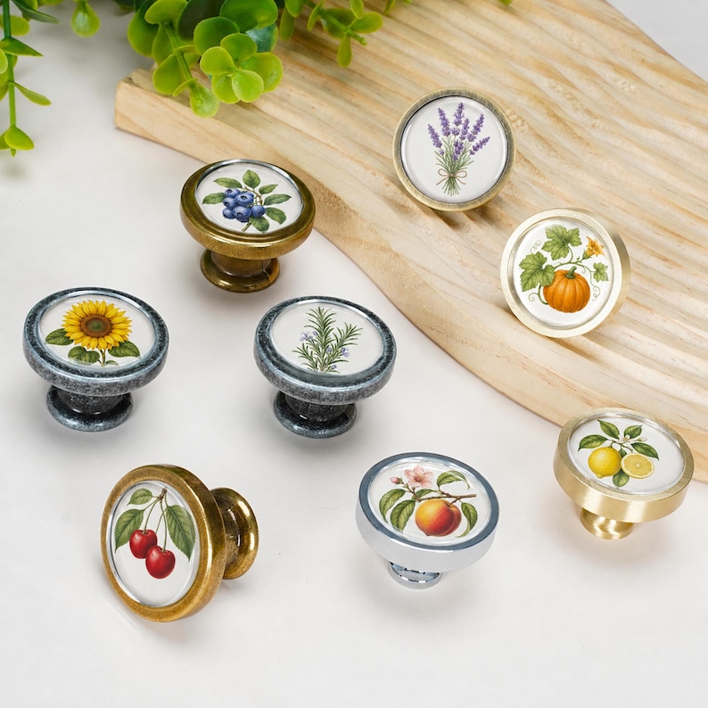 May include: A collection of seven round cabinet knobs featuring botanical and fruit illustrations. The designs include lavender, a pumpkin, blueberries, a sunflower, rosemary, cherries, a peach, and lemons, set in metallic finishes of gold, silver, and bronze.