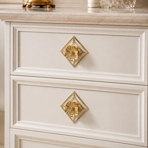 May include: A white cabinet featuring two drawers, each equipped with a gold-toned metal pull. The pull consists of a central flower-shaped knob mounted within a diamond-shaped frame, providing a decorative accent against the flat drawer front.