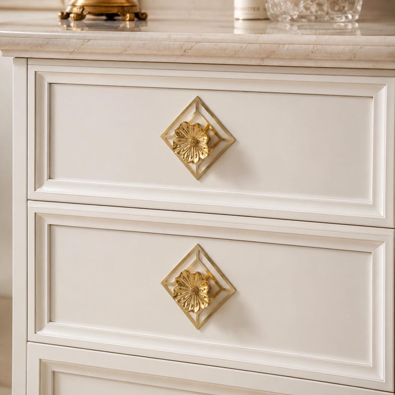 May include: A white cabinet featuring two drawers, each equipped with a gold-toned metal pull. The pull consists of a central flower-shaped knob mounted within a diamond-shaped frame, providing a decorative accent against the flat drawer front.