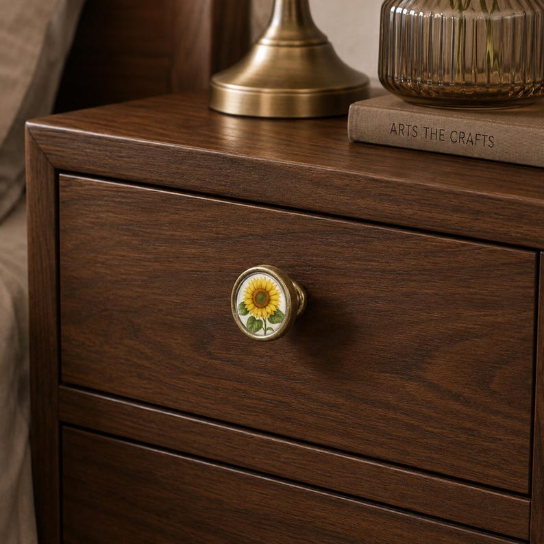 May include: A round cabinet knob with a brass-toned metal rim and a central circular inlay featuring a detailed illustration of a yellow sunflower with green leaves. The knob is mounted on a dark brown wooden drawer front with visible grain.
