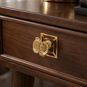 May include: A gold-toned metal drawer pull featuring a three-dimensional orchid flower shape mounted on a square geometric backplate, attached to a dark brown wooden drawer front with a visible wood grain texture and a smooth finish.