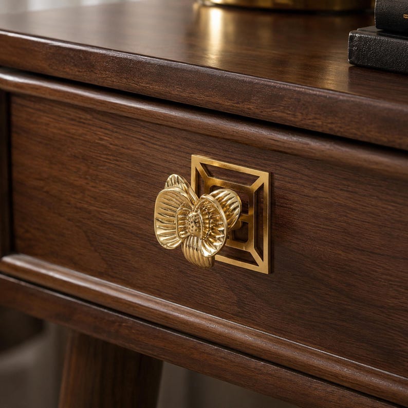May include: A gold-toned metal drawer pull featuring a three-dimensional orchid flower shape mounted on a square geometric backplate, attached to a dark brown wooden drawer front with a visible wood grain texture and a smooth finish.