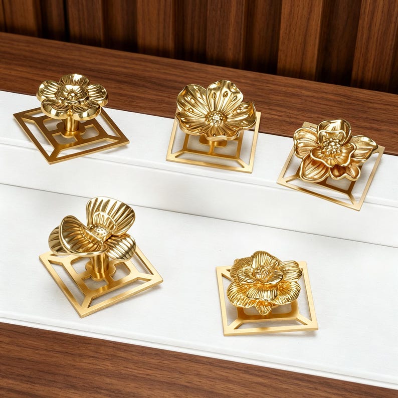 May include: Five gold-colored metal cabinet knobs, each featuring a three-dimensional flower design mounted on a flat, square, open-frame base. The metallic finish is reflective, highlighting the detailed petal textures and the central stamens.