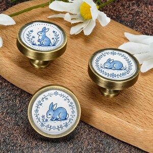 May include: Three round cabinet knobs with a brushed brass finish and a white ceramic center inlay. Each knob features a blue illustration of a rabbit surrounded by a delicate blue floral wreath. The designs show rabbits in different poses.