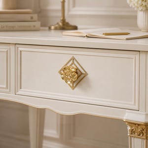 May include: A cream-colored wooden drawer featuring a central gold-toned metal knob shaped like a flower, mounted on a square geometric frame. The drawer front has a recessed rectangular panel design with gold trim along the bottom edge.