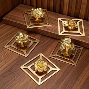 May include: Five gold-colored metal cabinet knobs, each featuring a three-dimensional flower design mounted on a square, open-frame backplate. The knobs have a polished, reflective finish and are arranged in a group on a wooden surface.