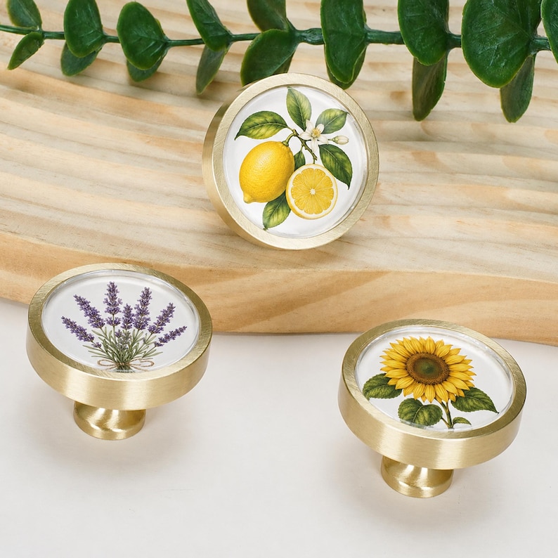May include: Three round cabinet knobs with a brushed gold metal finish. Each knob features a circular insert displaying a botanical illustration: one with a yellow lemon and leaves, one with purple lavender, and one with a yellow sunflower.