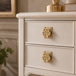 May include: A white wooden nightstand featuring two drawers, each equipped with a square gold-toned metal pull. The pulls are centered on the drawer fronts and feature a raised, detailed floral design with five petals set against a geometric frame.