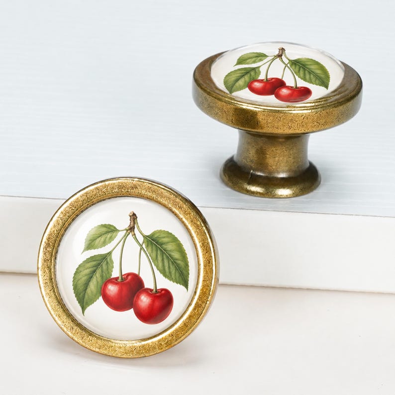 May include: Two round cabinet knobs featuring a brass-toned metal finish and a glass-covered insert. The insert displays a detailed botanical illustration of two bright red cherries with green leaves on a white background.
