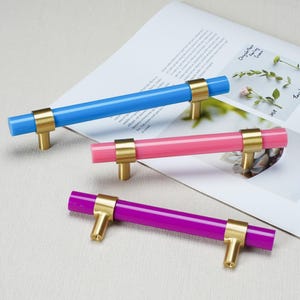 May include: Three cylindrical cabinet pulls with a smooth finish, each featuring a vibrant colored bar in blue, pink, or purple, secured by two brushed gold metal posts. The hardware is designed for mounting with visible screw holes at the base.