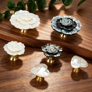 May include: A collection of six decorative cabinet knobs featuring floral and heart shapes. Four knobs are carved into layered rose designs in white and dark gray, while two heart-shaped knobs feature a white iridescent tiled pattern on brass bases.