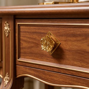 May include: A wooden drawer front featuring a polished gold-tone metal knob shaped like a flower, mounted on a square geometric backplate. The drawer is accented with thin gold-tone trim lines and vertical decorative gold-tone metal filigree.