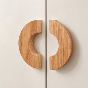 May include: Two semi-circular light brown oak wood cabinet handles mounted on a pair of off-white cabinet doors. The handles feature a visible natural wood grain pattern and are positioned to form a circular shape when the doors are closed.