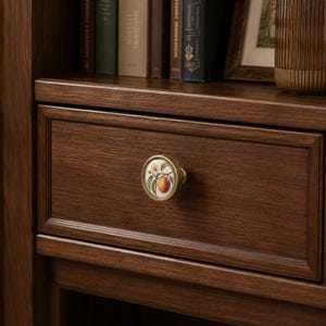 May include: A round drawer knob with a brass-toned metal rim and a white ceramic centre featuring a botanical illustration of a peach with green leaves and a small flower, mounted on a dark brown wooden drawer front with a frame detail.