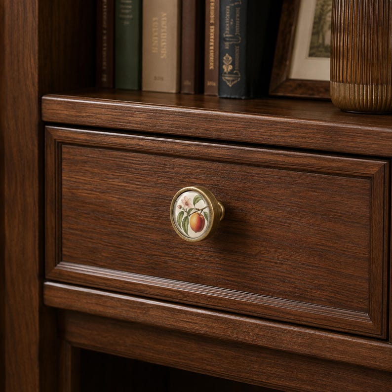 May include: A round drawer knob with a brass-toned metal rim and a white ceramic centre featuring a botanical illustration of a peach with green leaves and a small flower, mounted on a dark brown wooden drawer front with a frame detail.