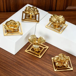 May include: Five gold-colored metal cabinet knobs, each featuring a three-dimensional flower shape mounted on a square, open-frame base. The metallic finish is polished and reflective, showcasing detailed petal textures and geometric square backplates.