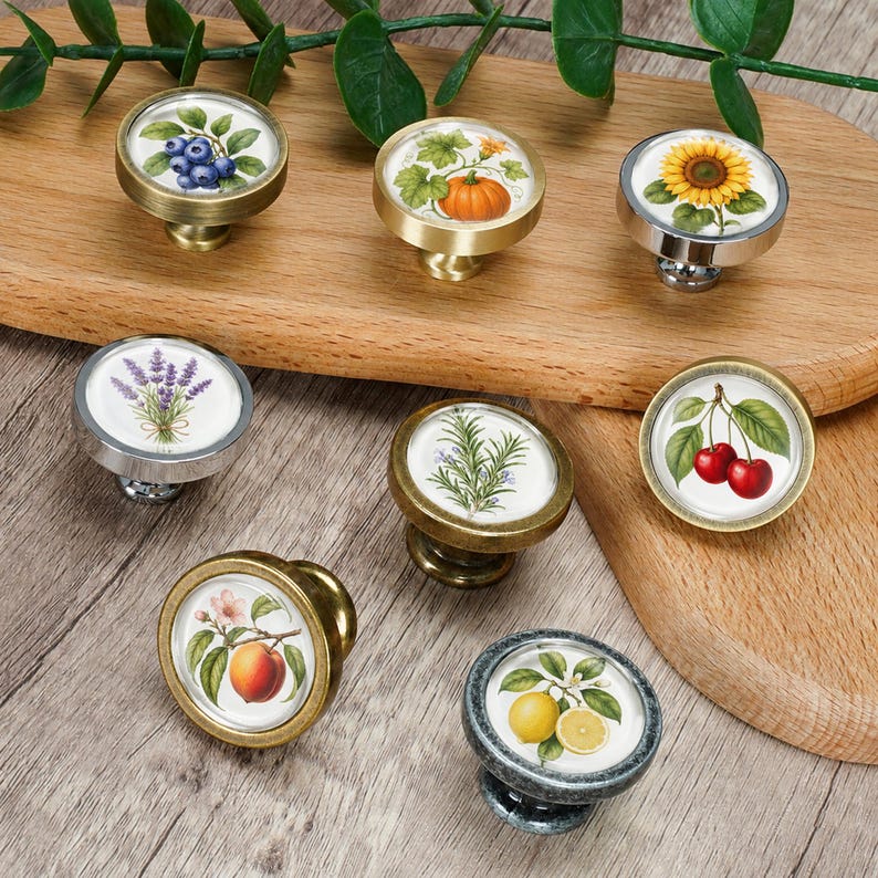 May include: A set of eight round cabinet knobs featuring botanical and fruit illustrations including blueberries, a pumpkin, a sunflower, lavender, rosemary, cherries, a peach, and lemons, set in metallic gold, silver, and bronze finishes.