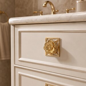 May include: A square cabinet knob featuring a gold-toned, three-dimensional floral design centered on a geometric frame. The knob is mounted on a cream-colored cabinet drawer with raised panel detailing, positioned below a marble countertop.