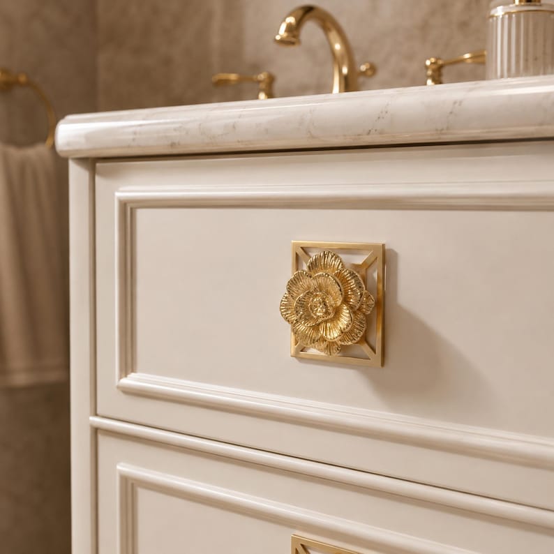 May include: A square cabinet knob featuring a gold-toned, three-dimensional floral design centered on a geometric frame. The knob is mounted on a cream-colored cabinet drawer with raised panel detailing, positioned below a marble countertop.
