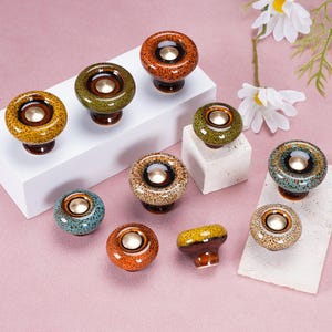 Puede incluir: Una colección de pomos redondos de cerámica para armarios con esmaltes moteados y multicolores en tonos naranja, verde, azul y marrón. Cada pomo tiene un acabado liso y brillante con una pieza central circular de metal dorado.