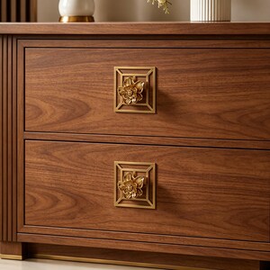 May include: A wooden nightstand with two drawers featuring dark brown wood grain. Each drawer is equipped with a square, gold-toned metal pull plate that frames a three-dimensional, sculpted gold flower knob centered on the drawer front.