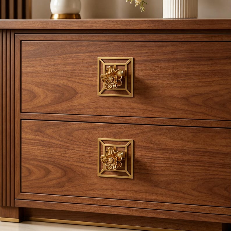 May include: A wooden nightstand with two drawers featuring dark brown wood grain. Each drawer is equipped with a square, gold-toned metal pull plate that frames a three-dimensional, sculpted gold flower knob centered on the drawer front.