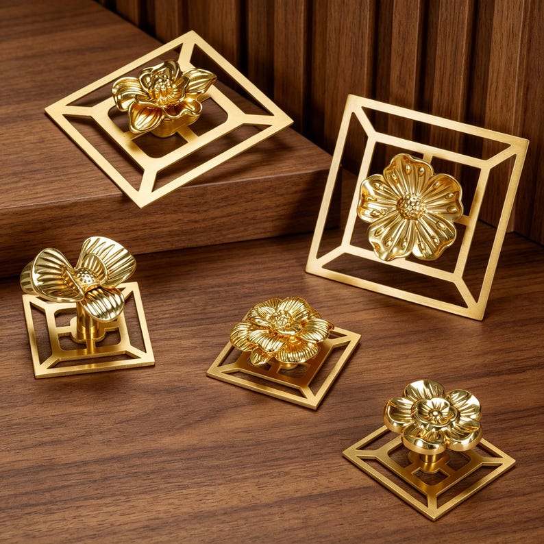 May include: A collection of five gold-toned metal cabinet knobs, each featuring a three-dimensional floral design centered on a square, open-frame geometric base. The flowers exhibit varied petal shapes and textures, all finished in a polished metallic gold.