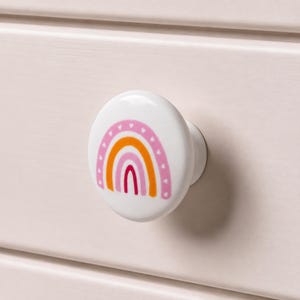 Puede incluir: Un pomo de cajón redondo de cerámica blanca con un estampado gráfico colorido de un arcoíris. El diseño del arcoíris consta de arcos concéntricos en tonos rosa, naranja y rojo, acentuados con pequeñas formas de corazón blancas en el borde.