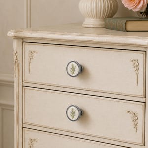 May include: A cream-coloured wooden chest of drawers featuring distressed edges and ornate carved floral corner details. Each drawer is fitted with a round ceramic knob decorated with a green botanical illustration of a rosemary sprig.