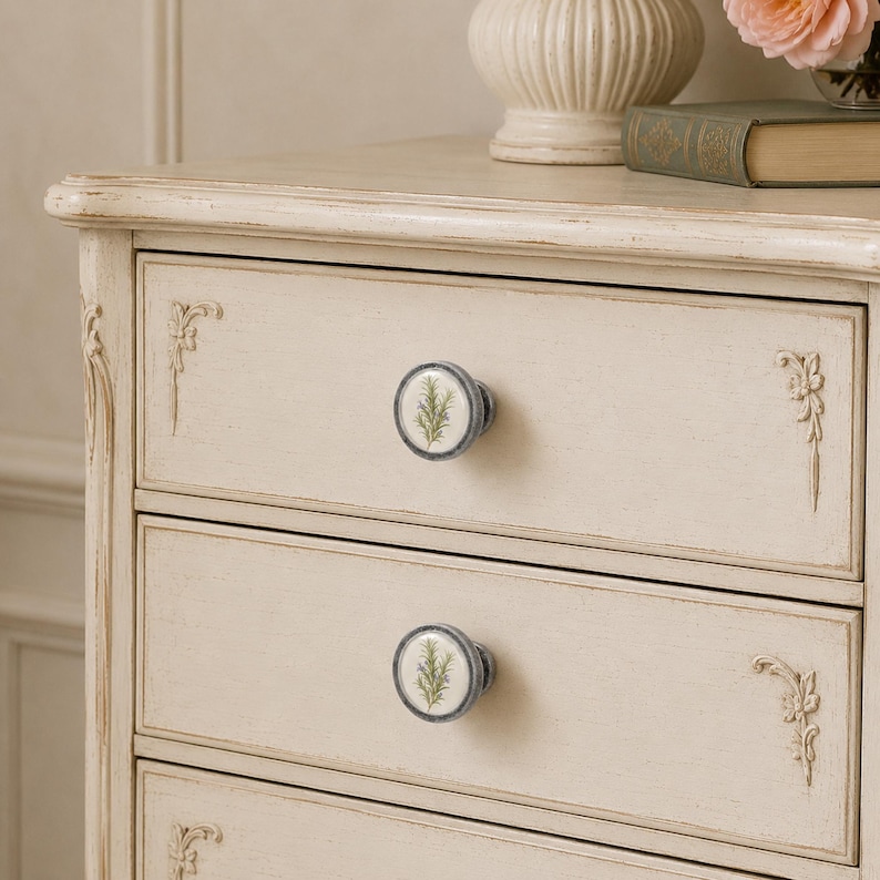 May include: A cream-coloured wooden chest of drawers featuring distressed edges and ornate carved floral corner details. Each drawer is fitted with a round ceramic knob decorated with a green botanical illustration of a rosemary sprig.