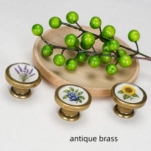 May include: Three round antique brass cabinet knobs with white ceramic inserts featuring botanical illustrations of lavender, blueberries, and a sunflower. A wooden bowl holds a green berry branch above the knobs. Text reads antique brass.