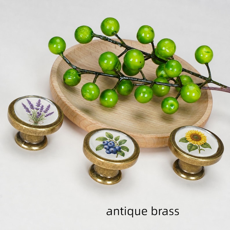 May include: Three round antique brass cabinet knobs with white ceramic inserts featuring botanical illustrations of lavender, blueberries, and a sunflower. A wooden bowl holds a green berry branch above the knobs. Text reads antique brass.