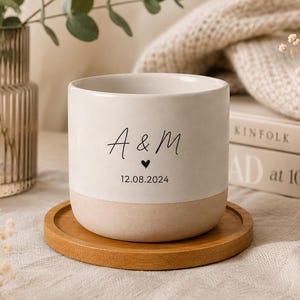 Puede incluir: Una taza cilíndrica de cerámica con diseño bicolor que presenta una sección superior en color crema mate y una base en tono beige natural. El frente muestra el texto negro A & M, un pequeño símbolo de corazón y la fecha 12.08.2024 sobre un posavasos.