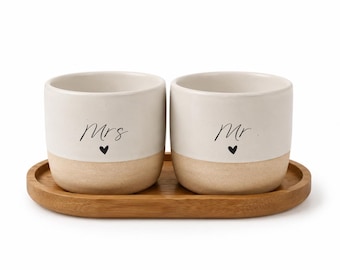 Può includere: Due tazze cilindriche in ceramica dal design bicolore, con una parte superiore bianco sporco opaco e una base color beige naturale. Ogni tazza presenta la scritta nera "Mrs" o "Mr" sopra un piccolo cuore nero, poggiate su un vassoio ovale in legno.
