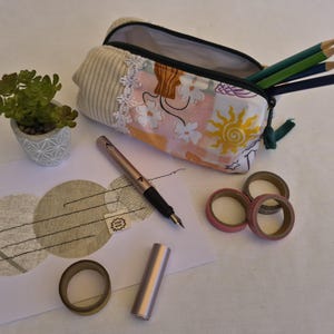 Könnte beinhalten: Ein Reißverschluss-Etui mit einer Seite aus beigem Rippstoff und einer Seite mit Blumenmuster und Sonnengrafik. Es enthält drei Buntstifte. Daneben liegen ein Füllfederhalter, eine kleine Sukkulente in einem Topf und vier Klebebandrollen.