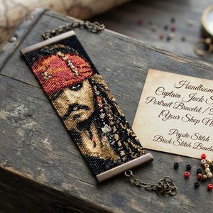 May include: A rectangular beaded bracelet featuring a portrait of Captain Jack Sparrow with a red headscarf, black hair, and beard. The piece includes metal end bars and a chain clasp. A card reads: Handloomed Captain Jack Sparrow Portrait Bracelet.