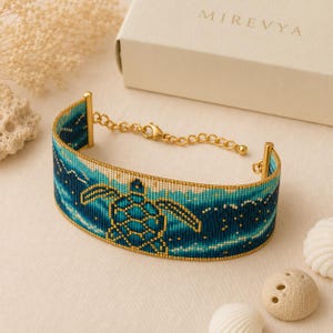 May include: A wide beaded bracelet featuring a sea turtle motif in shades of teal, navy blue, and gold. The design depicts a turtle swimming through ocean waves. It includes a gold-toned metal chain extender with a lobster claw clasp.