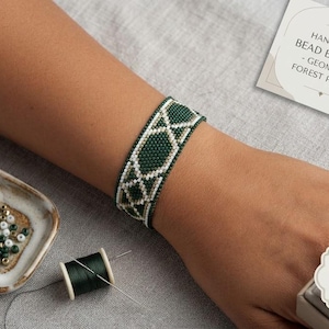 Puede incluir: Una pulsera de cuentas tejida a mano con un patrón geométrico de bosque en cuentas de color verde oscuro, blanco y oro metálico. La pulsera muestra un diseño de celosía repetitivo. Una pequeña tarjeta dice: HANDWOVEN BEAD BRACELET - GEOMETRIC FOREST PATTERN.