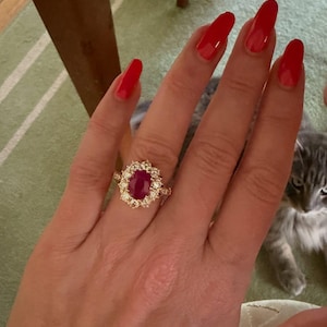 Op de afbeelding: Een gouden ring met een centrale ovaalvormige dieprode edelsteen omringd door een halo van twaalf ronde witte stenen. De band is dun en metaalachtig en toont een klassiek clusterontwerp met een opvallende gekleurde steen in het midden.