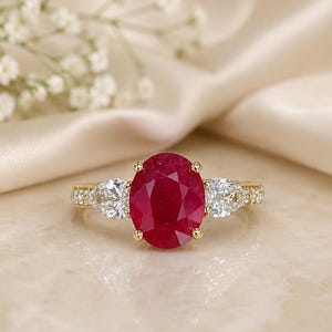 Op de afbeelding: Een gouden ring met een grote ovaal geslepen dieprode robijn in het midden, geflankeerd door twee peervormig geslepen witte diamanten aan weerszijden. De ringband is gemaakt van geelgoud en is bezet met kleine pavé-diamanten langs de schacht.