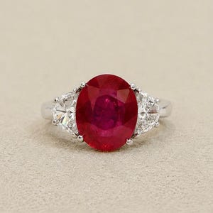Op de afbeelding: Een zilverkleurige ring met een grote, ovaal geslepen, dieprode robijn als centrale steen. De robijn wordt aan weerszijden geflankeerd door een heldere, driehoekig geslepen diamant, gezet in een gepolijste metalen band met zettingen.