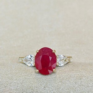 May include: A gold ring featuring a large oval-cut red ruby centre stone held by four prongs. The ruby is flanked by two pear-cut white diamonds on each side, set on a gold band that includes small pavé-set diamonds along the visible shank.