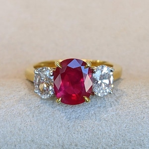 Könnte beinhalten: Ein Goldring mit einem großen, oval geschliffenen, tiefroten Rubin in der Mitte, der von vier Gelbgoldkrappen gehalten wird. Der Rubin ist auf beiden Seiten von einem kleineren, oval geschliffenen, klaren weißen Diamanten flankiert.