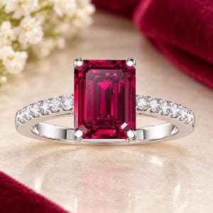 May include: A silver-toned ring featuring a large, rectangular emerald-cut deep red gemstone as the centre stone, held by four prongs. The band is crafted from a polished metallic material and is accented with a row of small, round, clear stones.